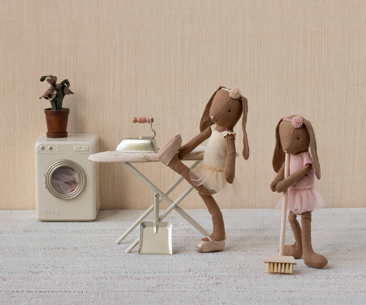 Two toy rabbit figures in a domestic setting with a washing machine, ironing board, and broom.